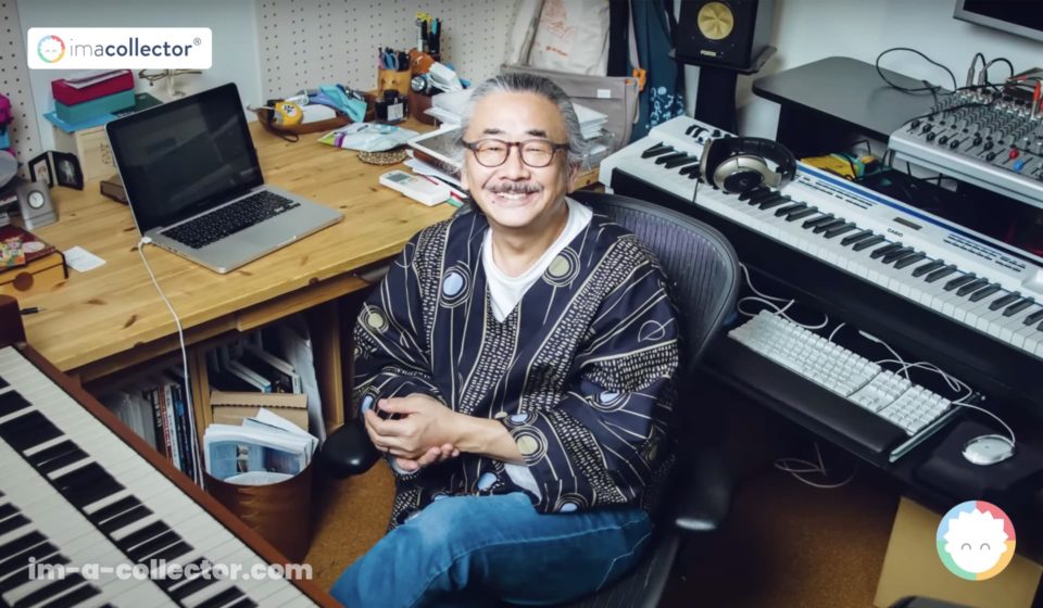 Nobuo Uematsu sourit au milieu de son matériel de création, entouré d’un ordinateur, d’un clavier et de son espace de travail.